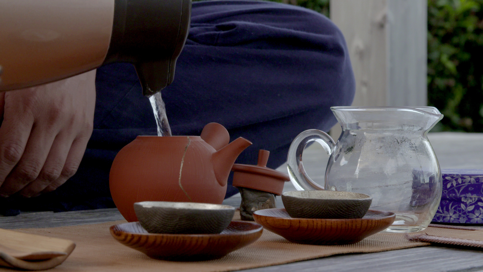 A close-up view of a person sitting on the floor pouring hot water from a kettle into a clay teapot placed on a mat. The scene includes two black cups on wooden saucers, a transparent glass pitcher with water, and a small colorful box in the background.