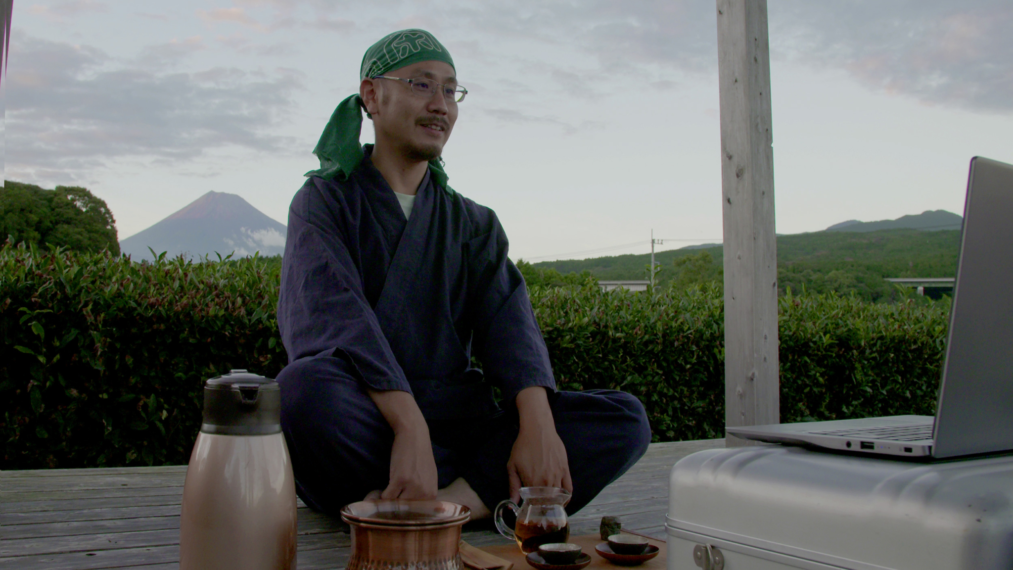 A person wearing traditional Japanese clothing and a green headscarf sits cross-legged on a wooden deck. They are pouring tea from a small pot into a cup, with a laptop and teapot nearby. Mount Fuji and a cloudy sky are visible in the background.