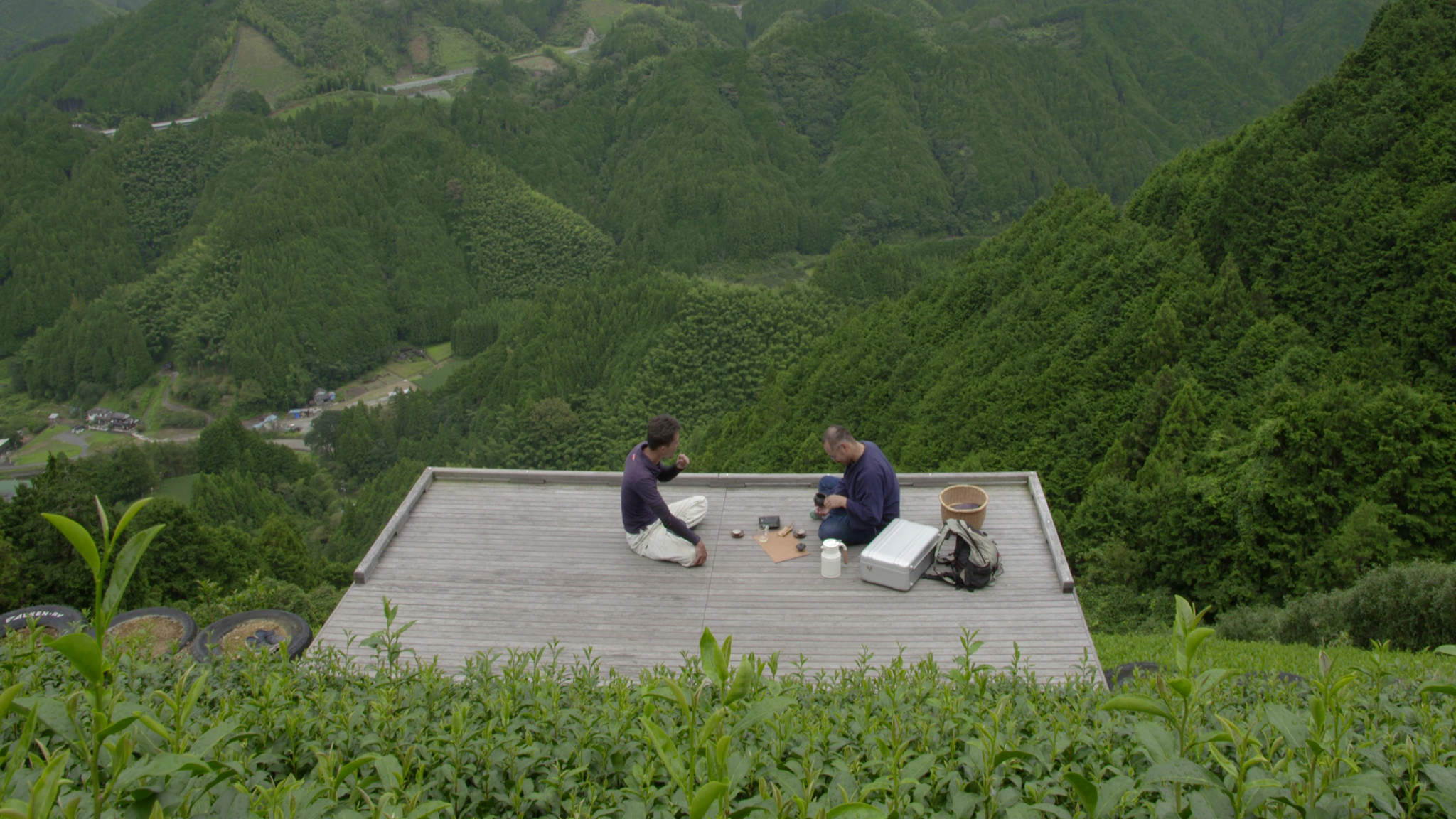 Two people sit on a wooden platform surrounded by lush green hills and mountains. The platform appears to be elevated from the ground and is surrounded by dense vegetation. They have some equipment and containers, and appear to be engaged in an activity together.