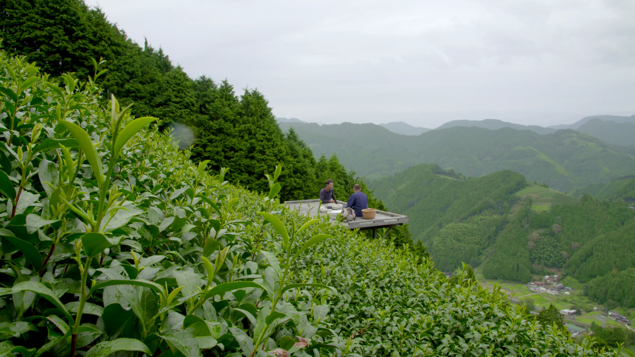 Two people are sitting on a wooden platform on a steep, green hillside surrounded by lush vegetation and overlooking a vast, mountainous landscape under a cloudy sky. The scene exudes tranquility and a strong connection to nature.