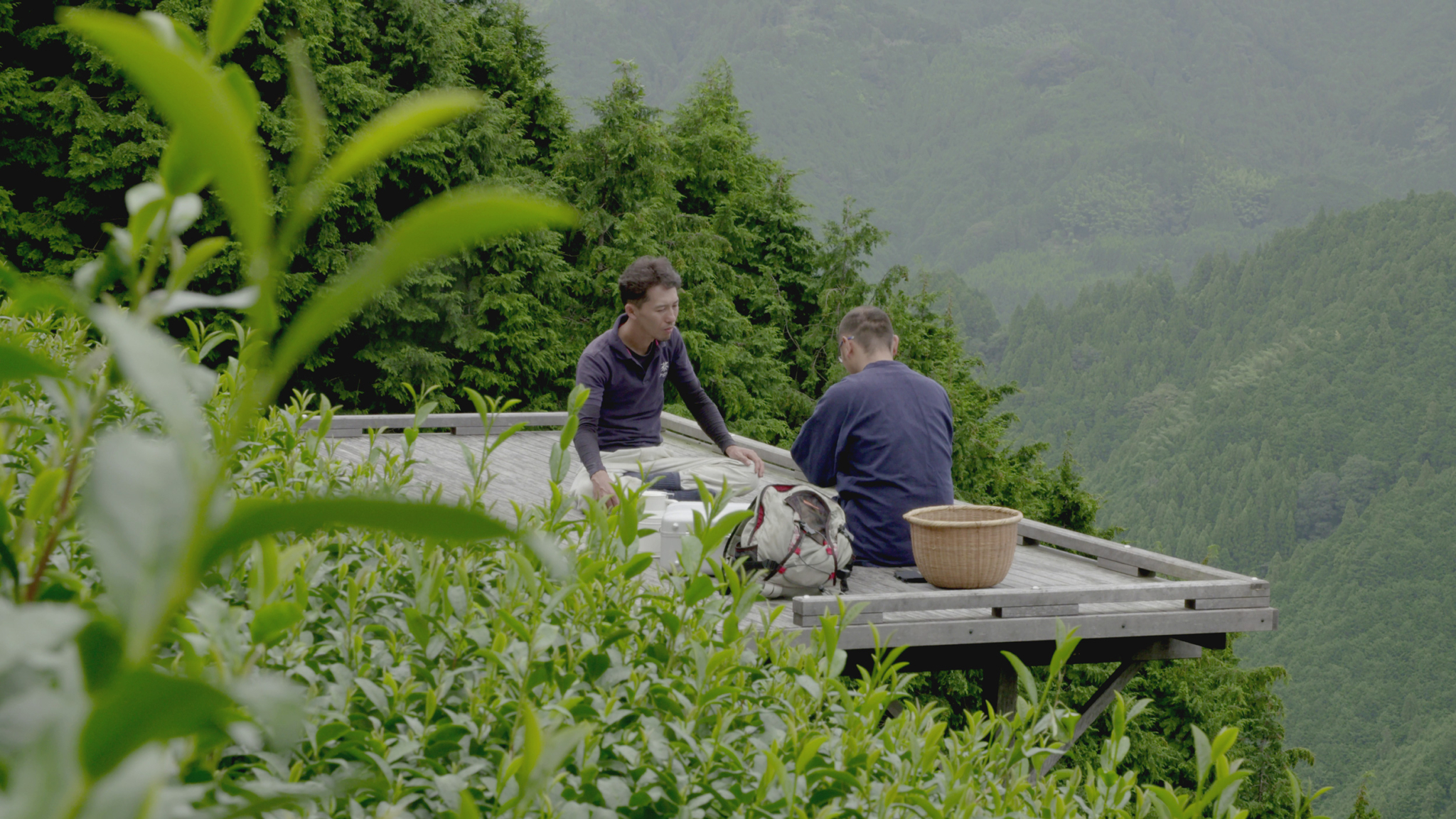 Two people sit on a wooden platform surrounded by lush greenery and hills. One person kneels, looking towards the other, who sits cross-legged. A wicker basket and a backpack are beside them. Dense green foliage and a mountainous landscape fill the background.