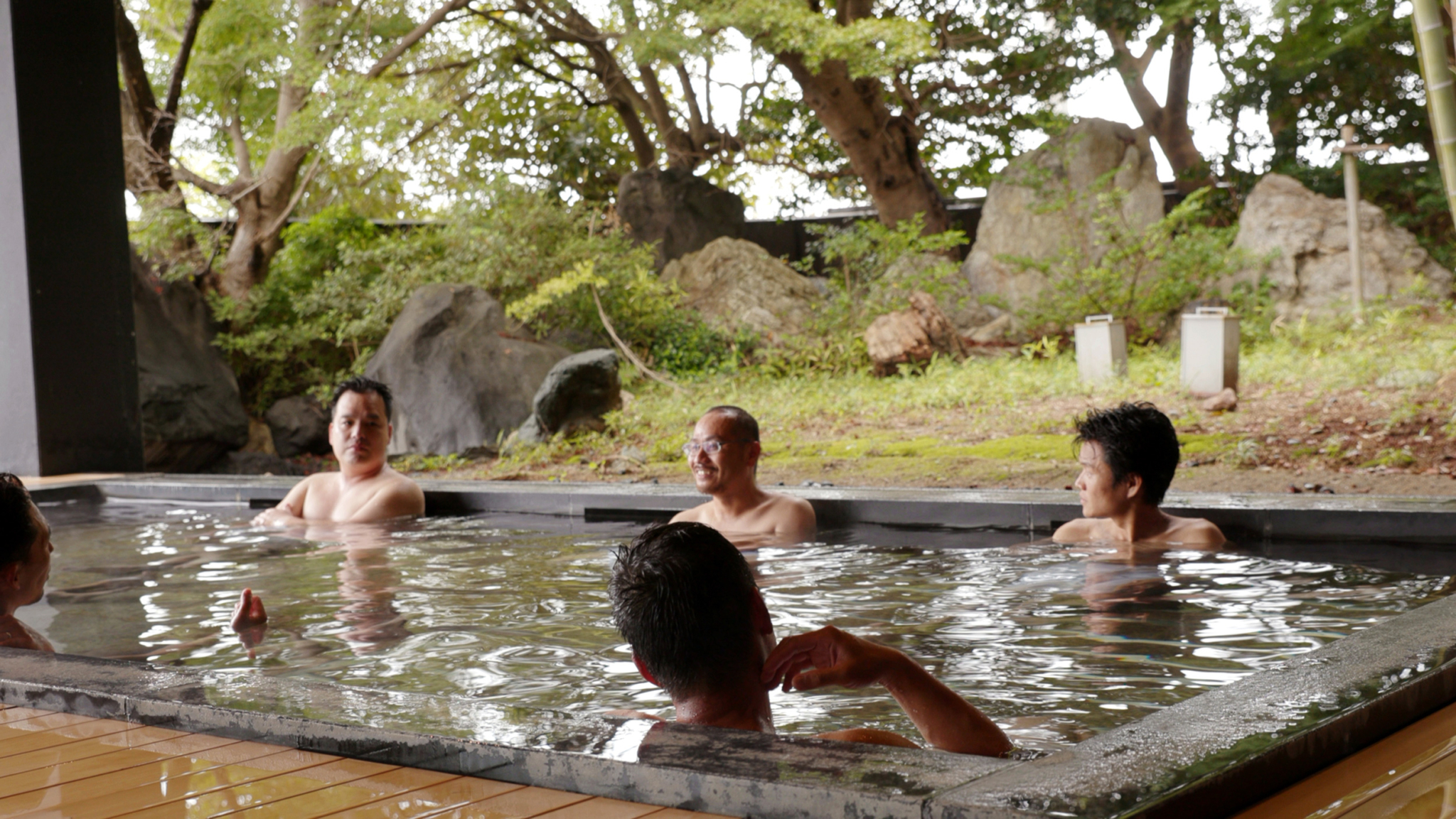 Four people are relaxing in an outdoor hot spring bath surrounded by lush greenery and rocks. They are seated in heated water that emits steam, looking relaxed and enjoying the serene natural environment. Trees and shrubs create a peaceful backdrop.