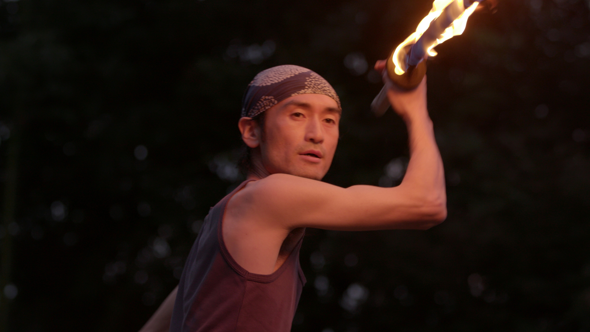A person wearing a dark tank top and a patterned headband is performing a fire dance. They are holding a flaming torch above their head, poised mid-action against a dark, blurred background.