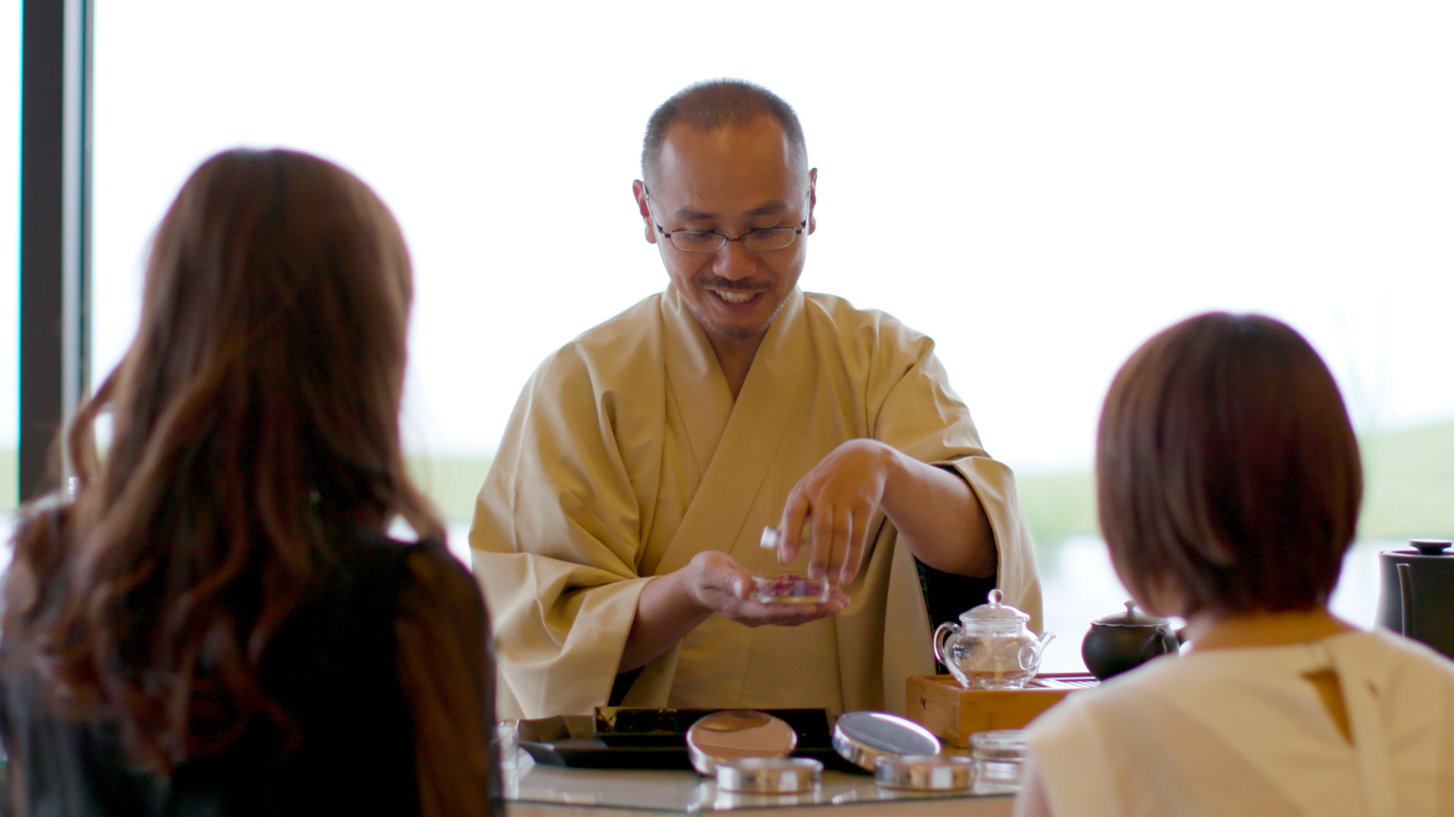 A man dressed in traditional Japanese attire is demonstrating a tea ceremony to two women seated in front of him. He is holding a small container, smiling, and appears to be in the middle of explaining part of the process. Teapots and other tea utensils are on the table.