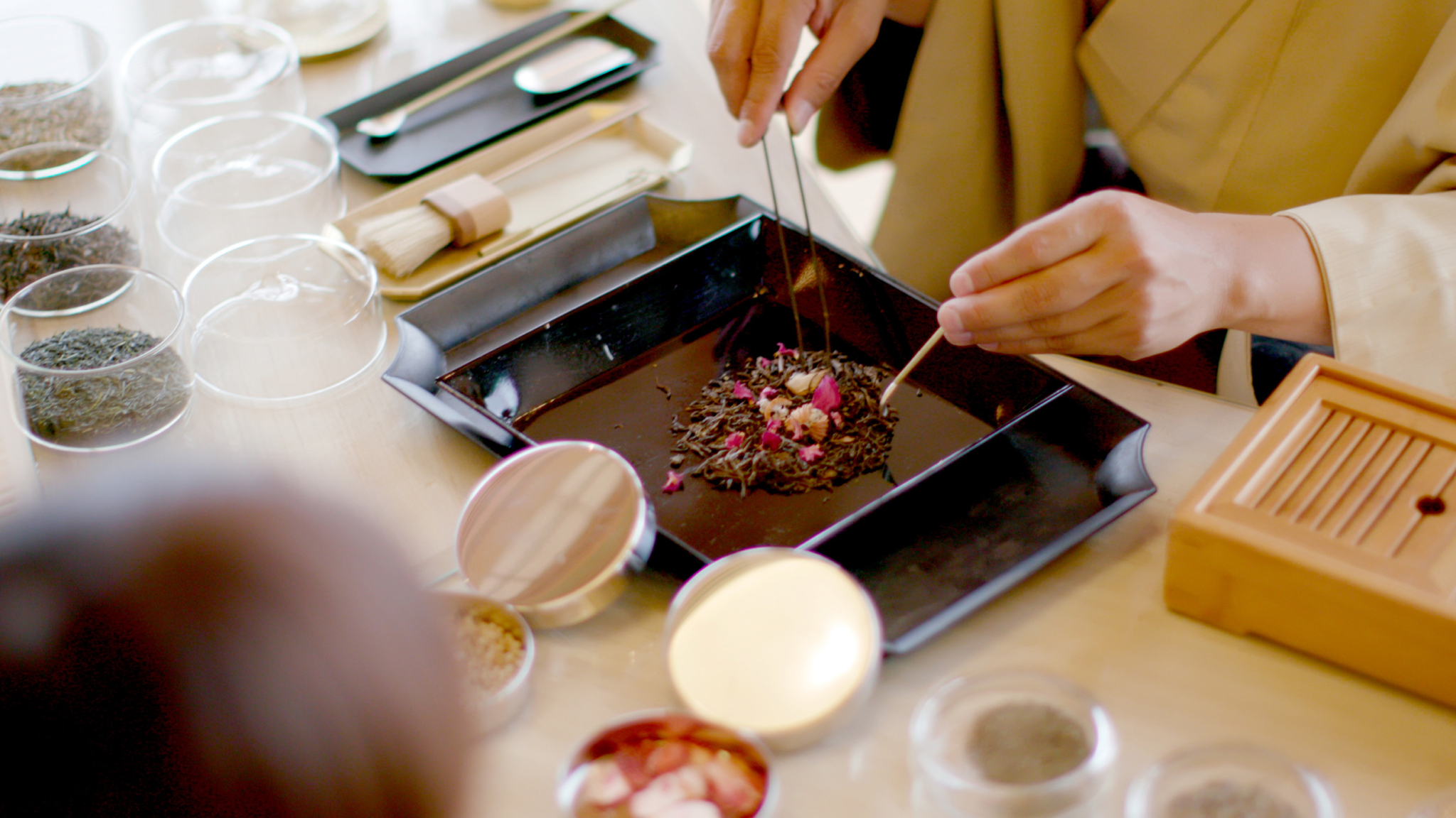 A person is carefully preparing a tray with various types of tea leaves and flowers. The setting includes several small containers holding different ingredients, utensils, and a wooden box on a clean, light-colored surface, suggesting a tea ceremony.
