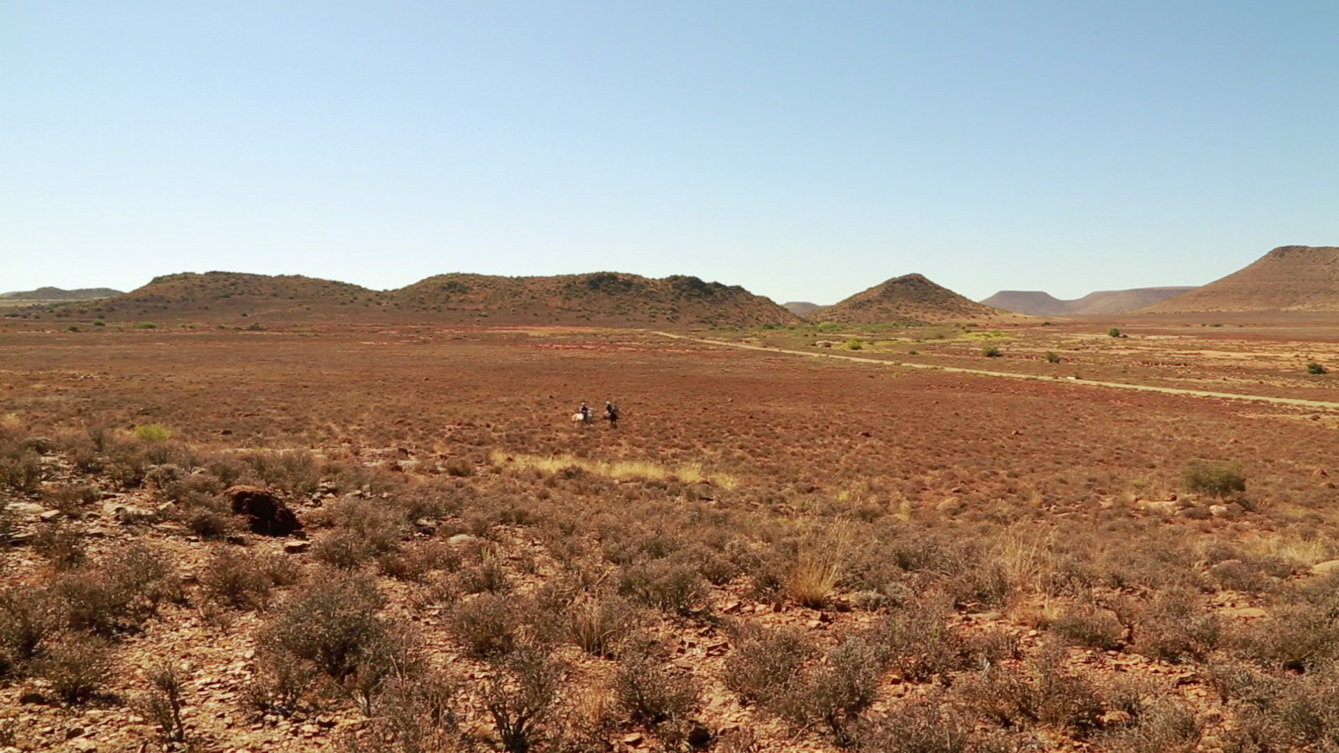 A vast, arid landscape with sparse vegetation and low, rolling hills under a clear blue sky. Two small figures can be seen in the distance, walking through the open expanse. The terrain is predominantly brown and dry, with patches of scrubby plants.
