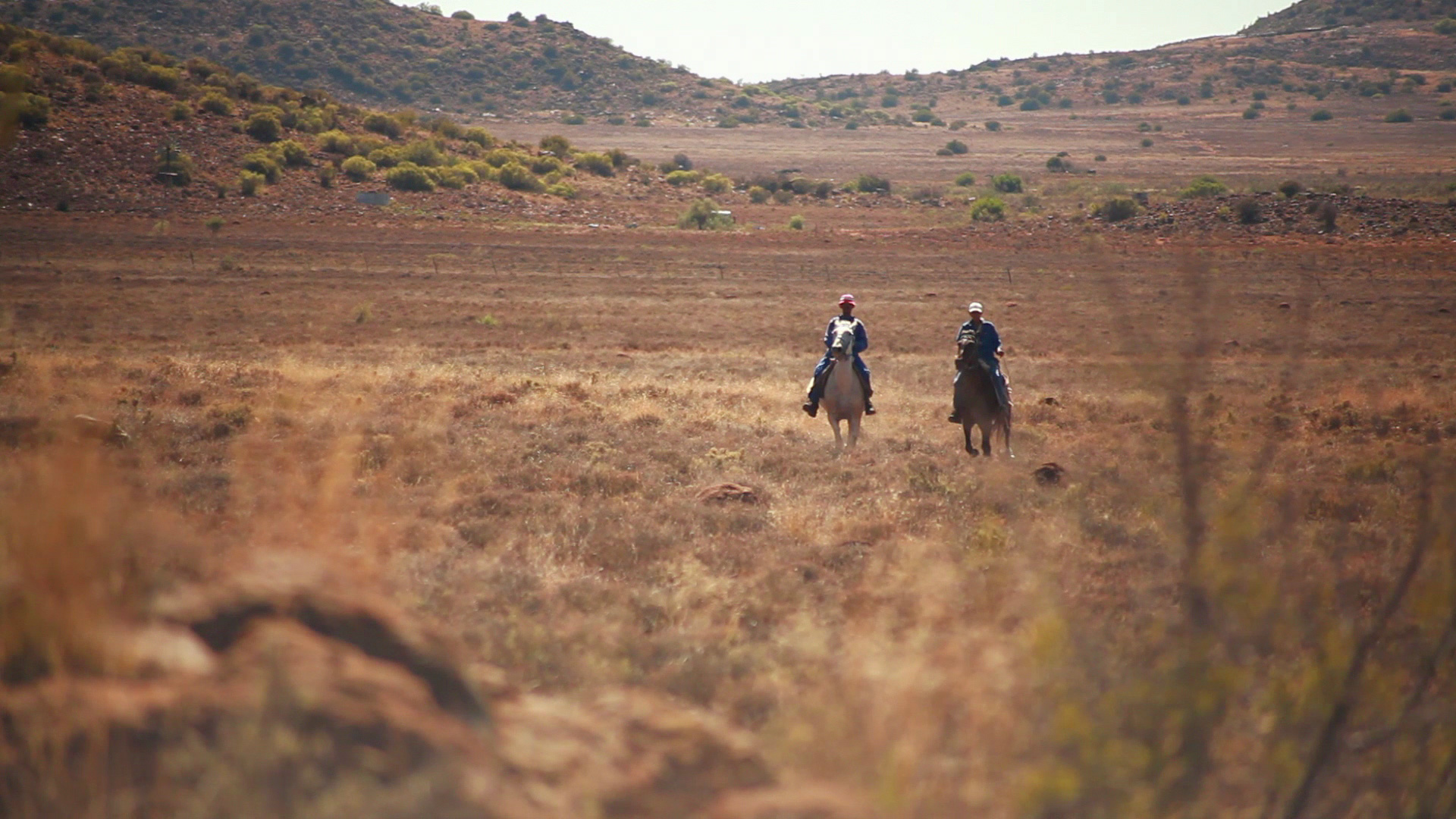 Two people riding horses walk through an expansive, dry, grassy landscape with rolling hills in the background. The terrain is arid with scattered bushes and earthy tones, suggesting a remote, open, and rugged environment.
