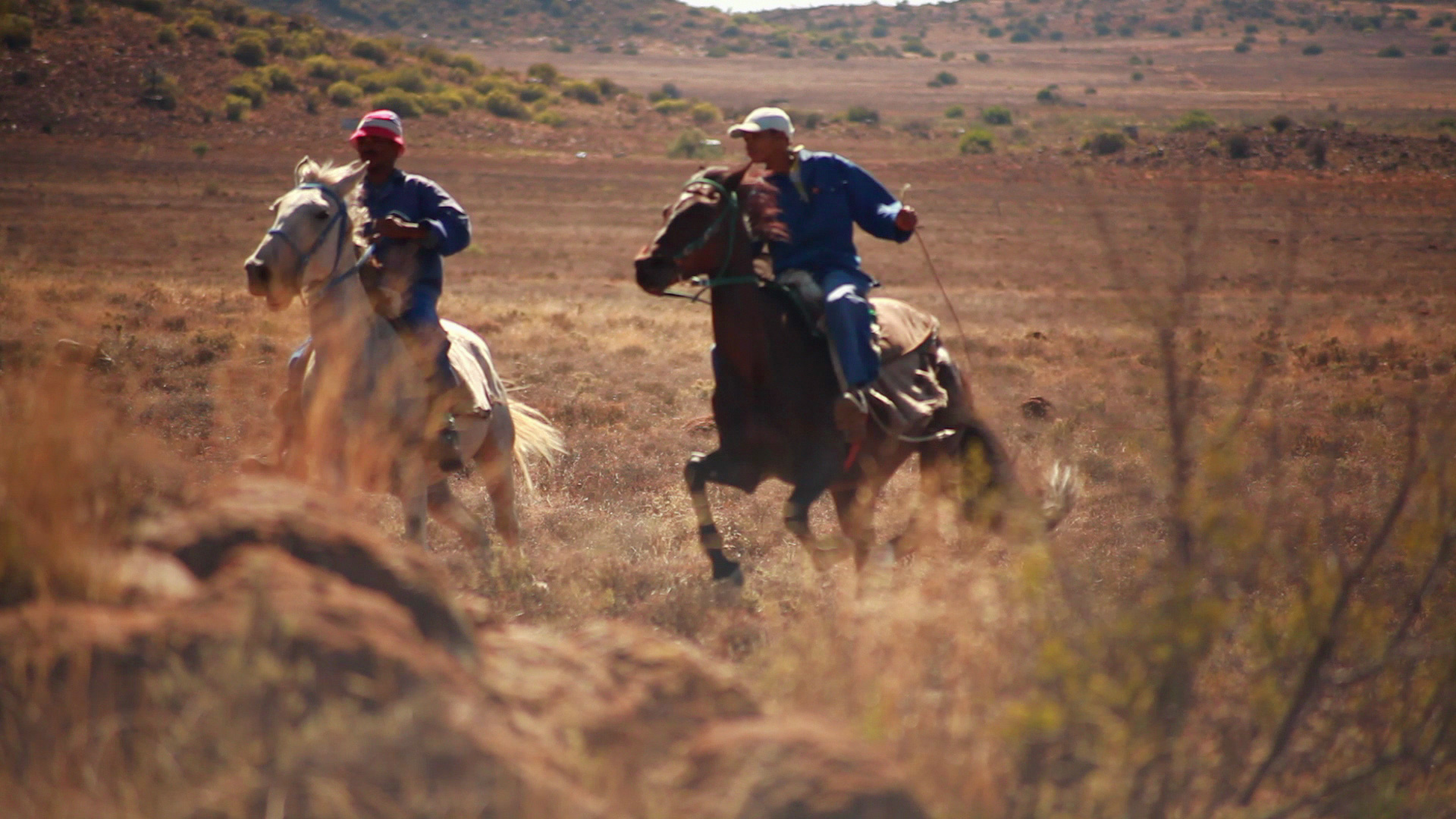 Two people riding horses across a dry, grassy landscape. The riders wear blue shirts and caps, one red and one beige. The terrain is rugged with sparse vegetation, and the background showcases a hilly area under a clear sky. The image captures a moment of motion.
