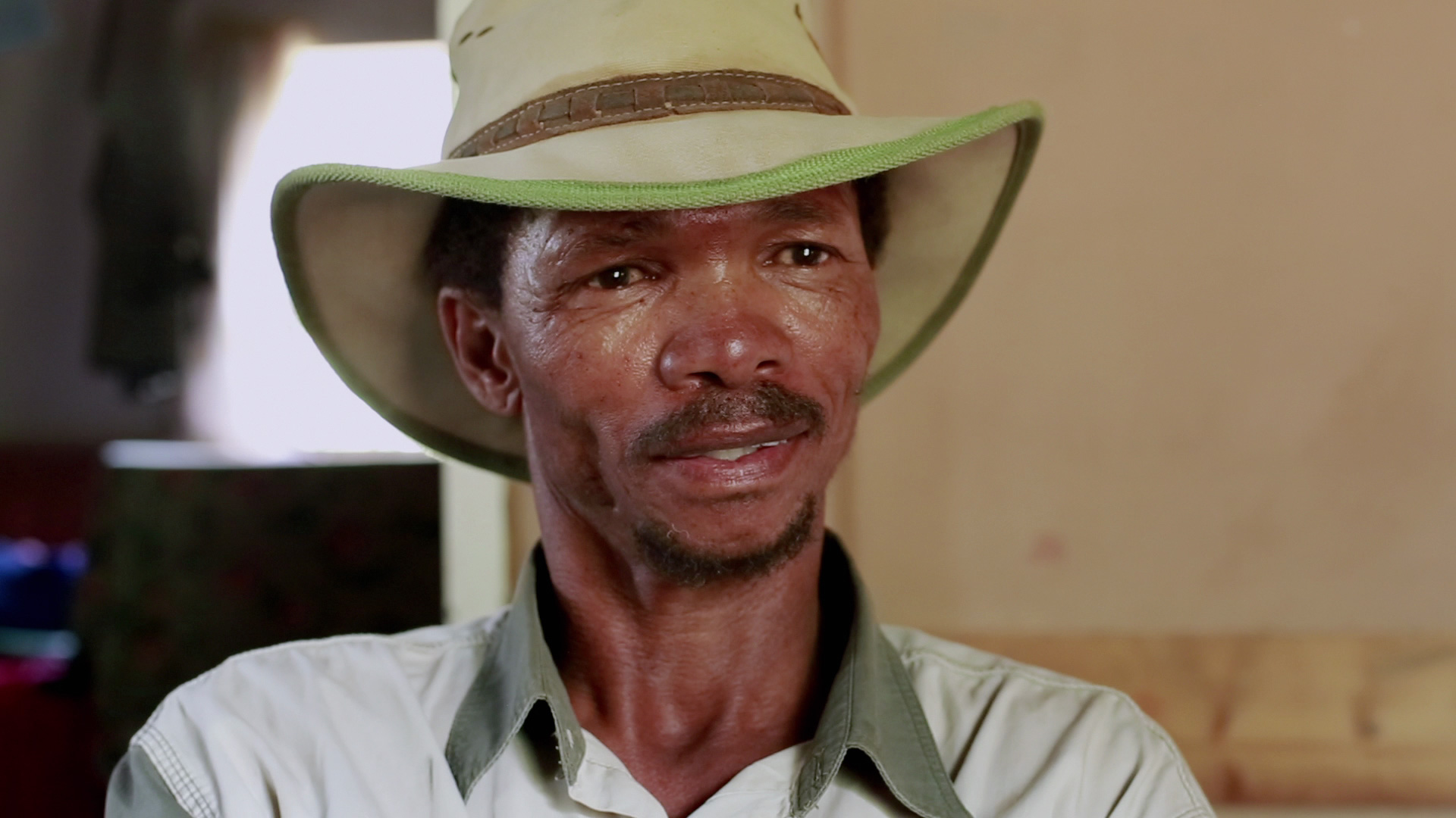 A middle-aged man with a weathered face is looking past the camera. He wears a wide-brimmed hat with a green rim and a light-colored shirt with a green collar. The background is mostly out of focus, featuring a partially lit room with warm tones.