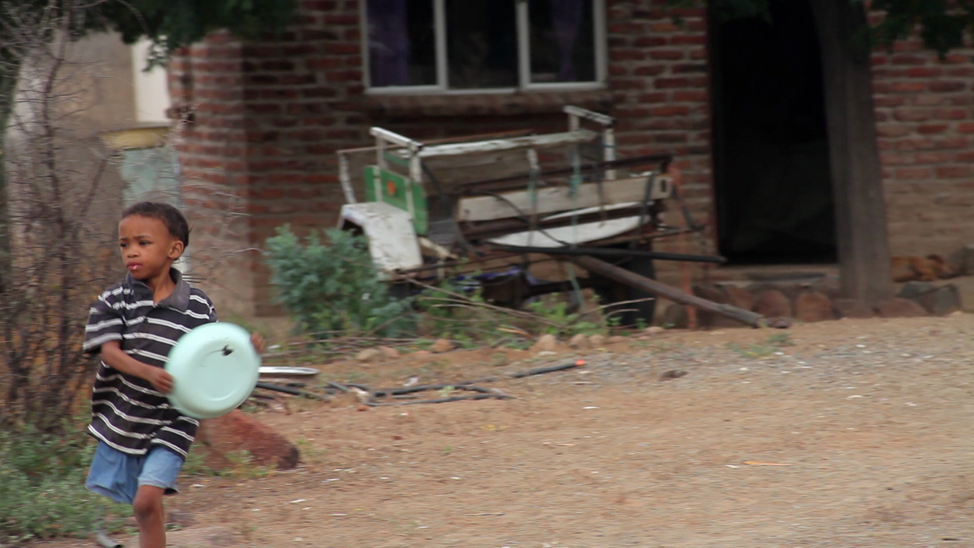 A young boy wearing a striped shirt and shorts walks across a dirt yard, holding a large container. Behind him, there is a brick building with a window and some discarded objects near the wall, including a metal cart.