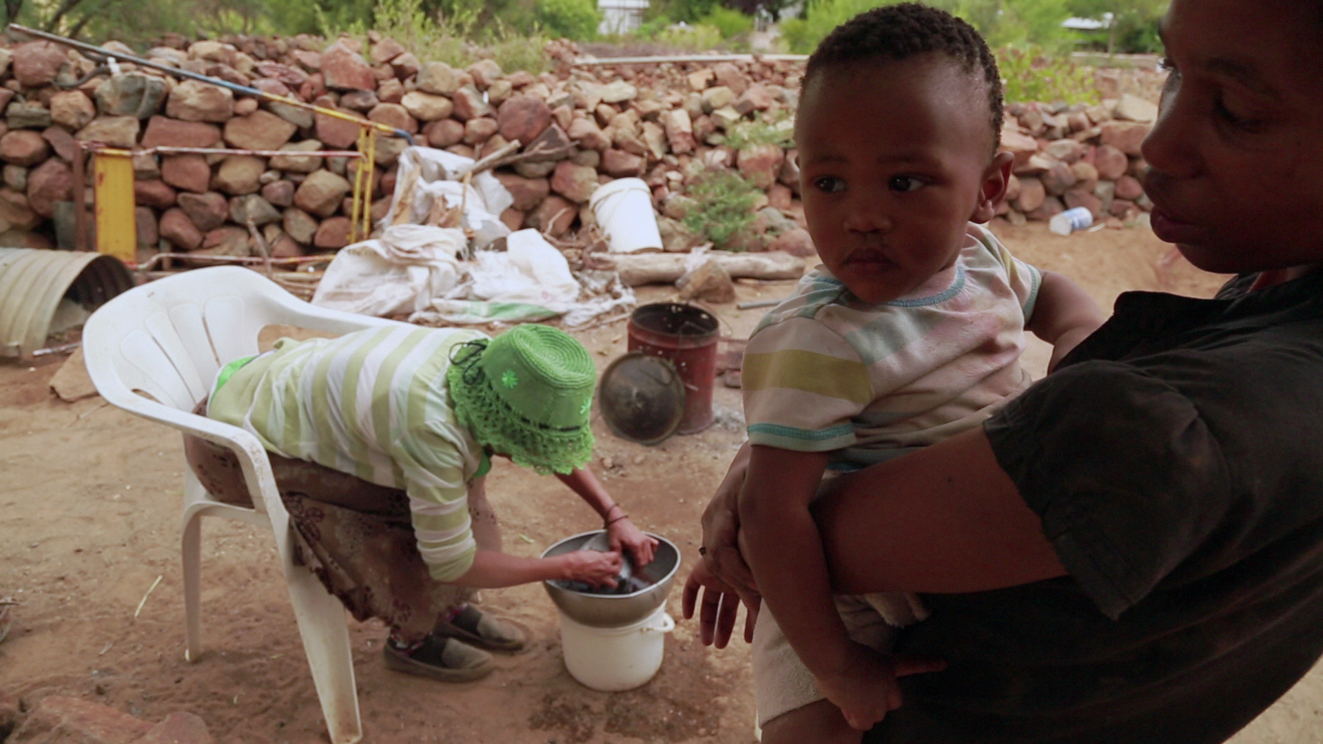 A woman wearing a green hat sits in a white plastic chair, washing clothes in a basin. In the foreground, another woman holds a young child. The scene is outdoors with a stone wall and various items scattered around.
