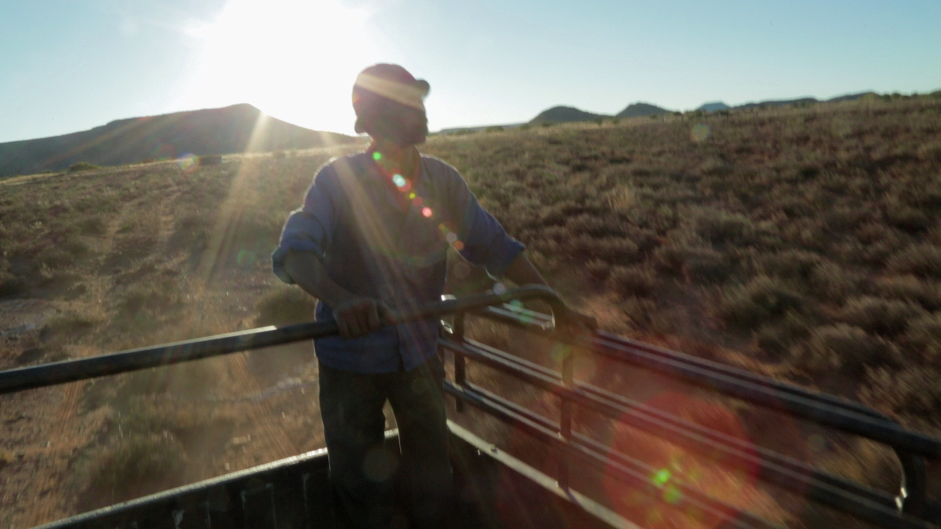 A person stands in the bed of a truck holding onto a railing, backlit by the sun. They are in an open, arid landscape with scattered vegetation and distant hills. The sun creates lens flare effects, casting rays and a halo around the person's silhouette.