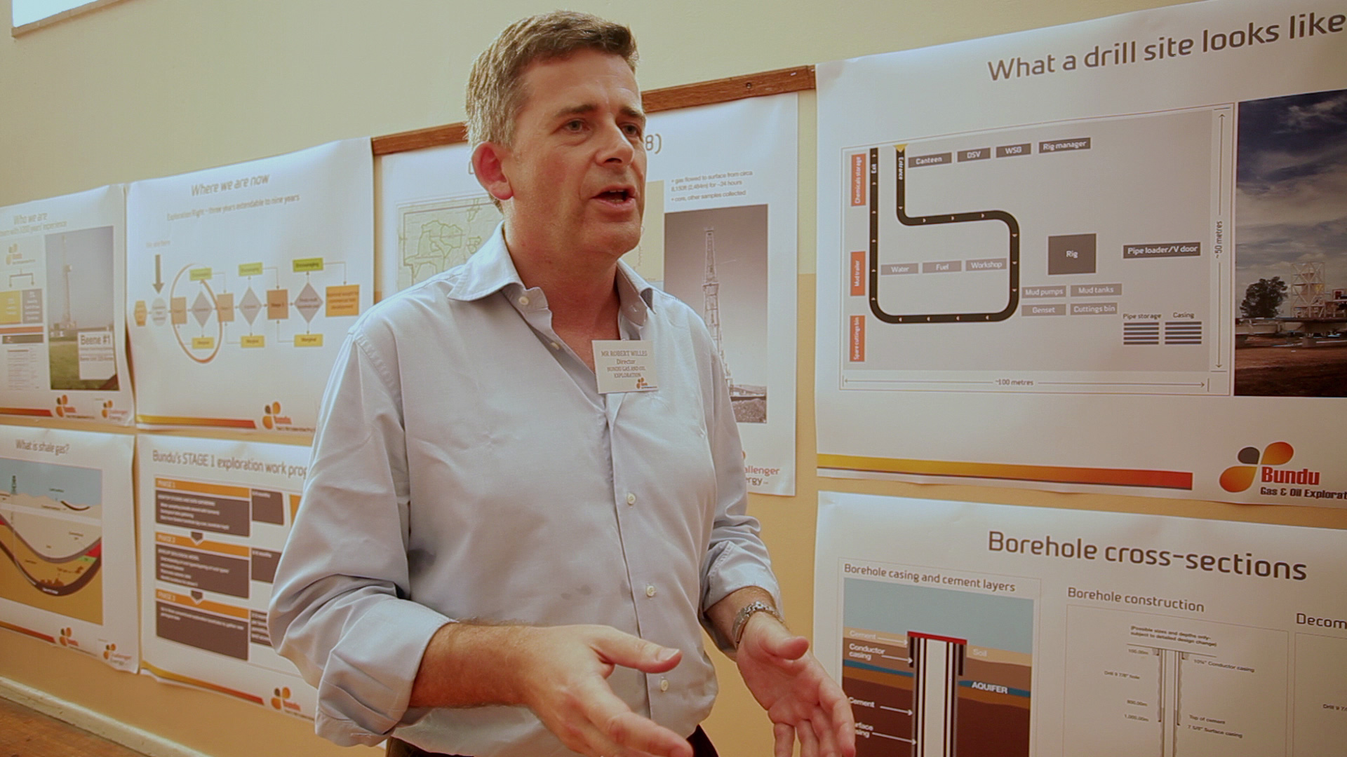 A man in a light blue shirt stands in front of a wall covered with various informational posters about drilling sites and borehole cross-sections. He appears to be gesturing as he speaks, with a name tag on his chest and a focused expression.