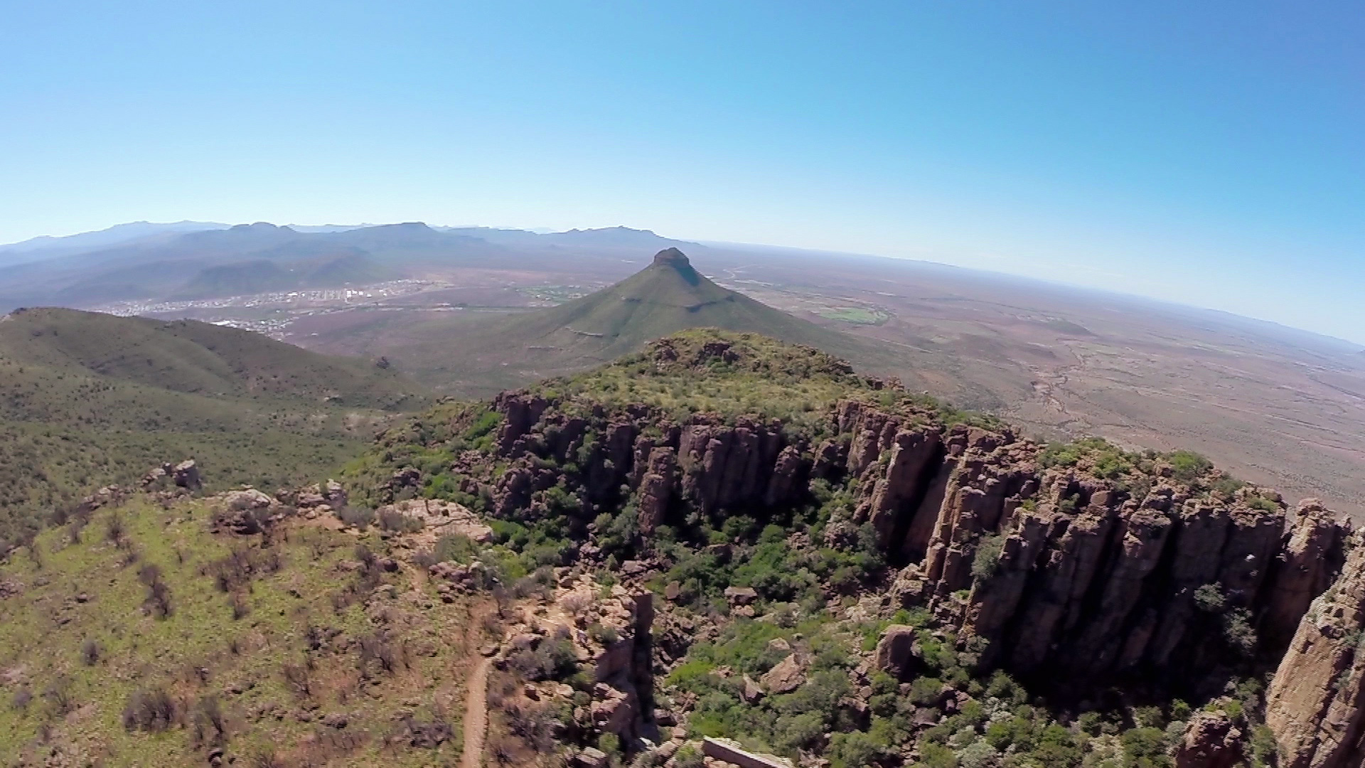 A panoramic landscape view shows a vast, green mountain range with a distinctive conical peak in the distance. The foreground features steep, rocky cliffs, and the horizon stretches under a clear blue sky, highlighting the expansive valley below.