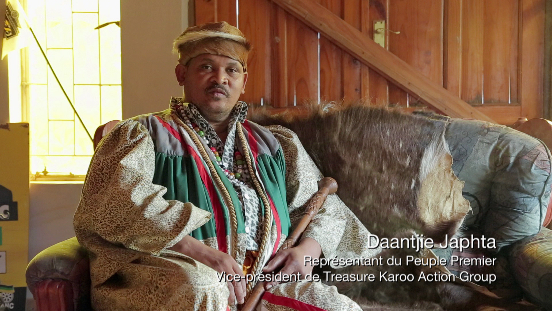 A person sits in a room decorated with wooden elements. They wear traditional attire, holding a staff, and have a neutral expression. Text on the image reads: "Daantjie Japhta, Représentant du Peuple Premier, Vice-président de Treasure Karoo Action Group.
