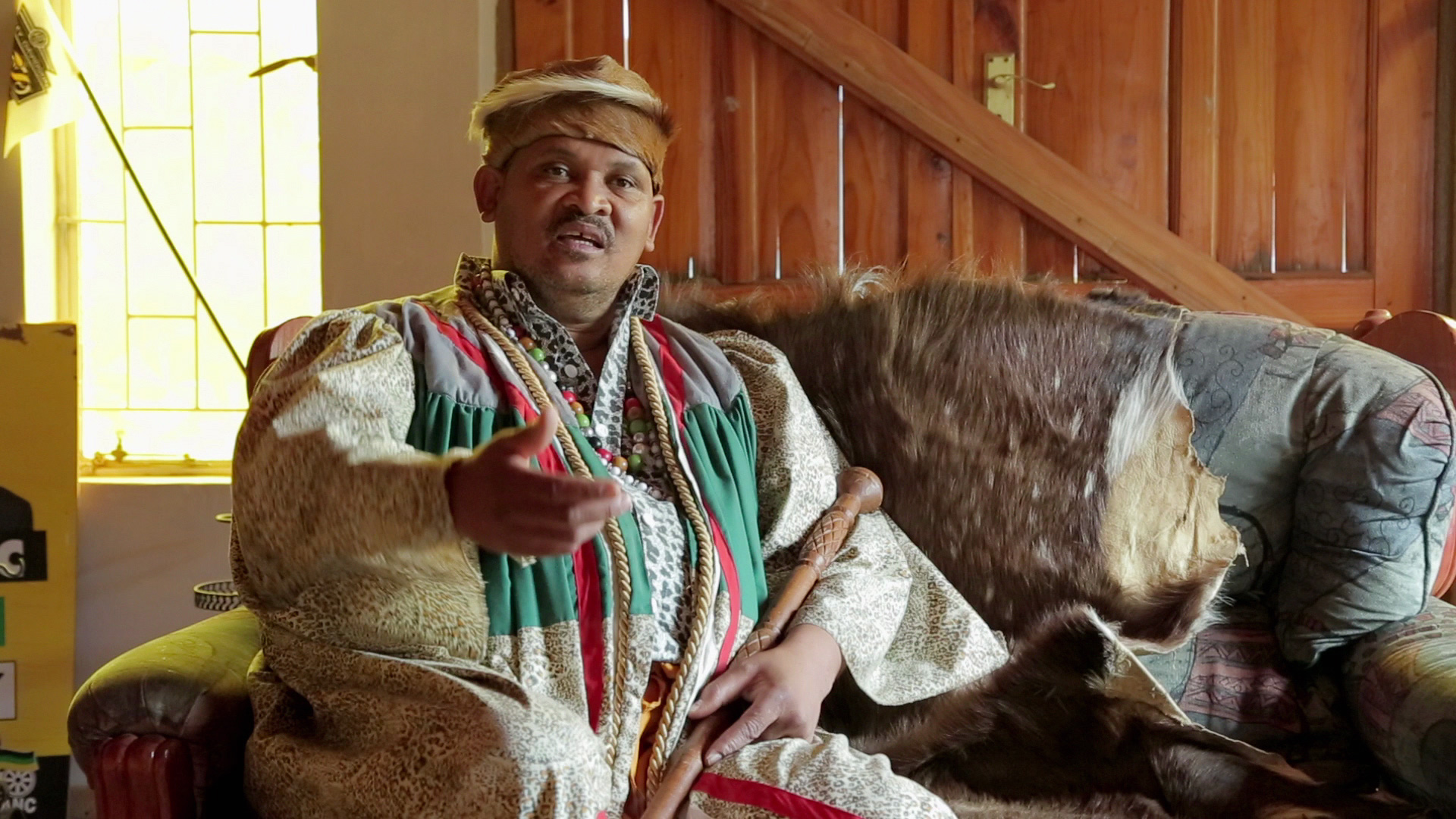 A person dressed in traditional attire sits on a patterned sofa in a room with wooden walls and a door. They are gesturing with one hand, while the other holds a carved wooden stick. Various fabric textures and patterns are visible around them.