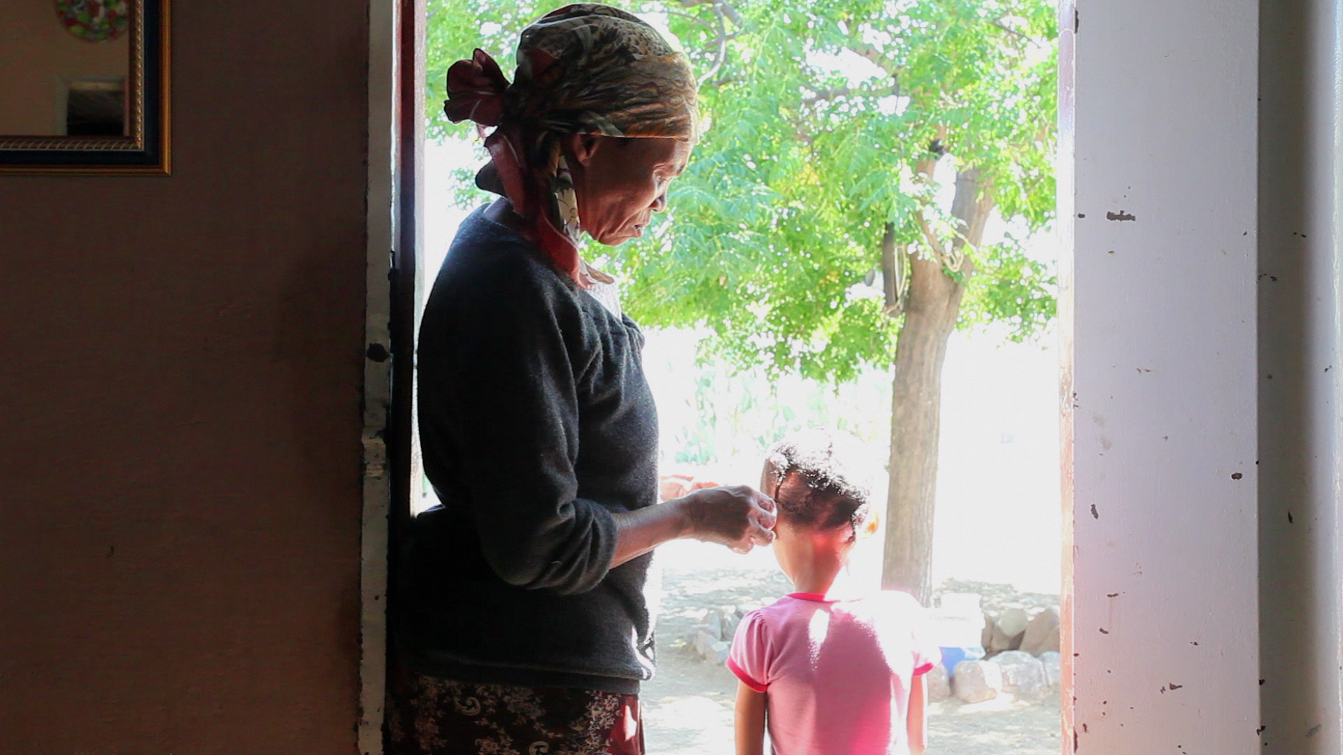 An elderly woman, wearing a headscarf, stands in a doorway with her hand on a young child's shoulder. The child, dressed in pink, faces away from the camera towards a bright outdoor scene with green trees. Sunlight filters through the doorway, illuminating them.