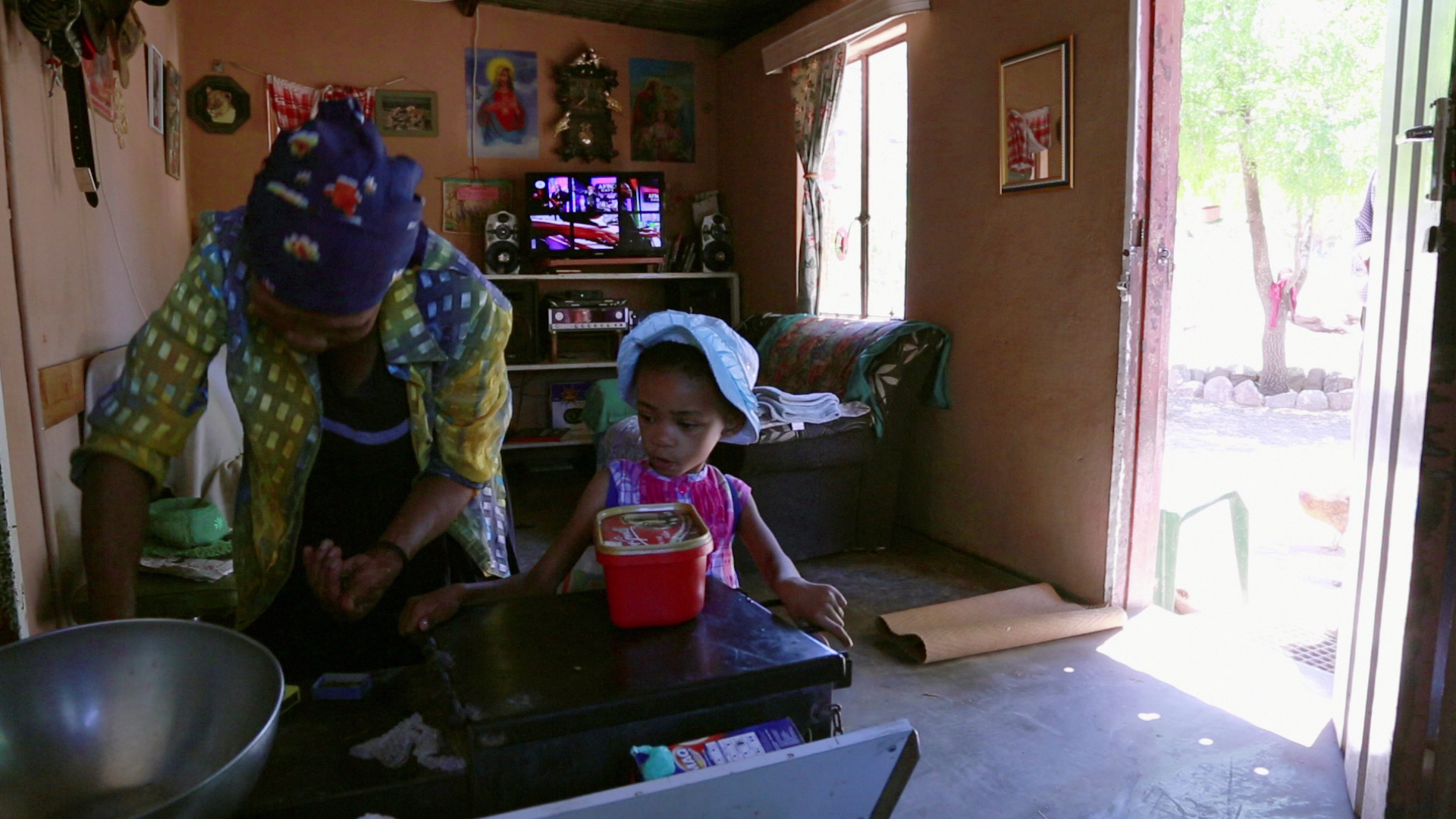 A woman in a headscarf and plaid shirt is working at a counter in a small, cozy room, while a young girl in a blue hat and purple dress stands beside her. The room is decorated with photos and artwork, and a TV is on in the background. Sunlight streams in through an open door.