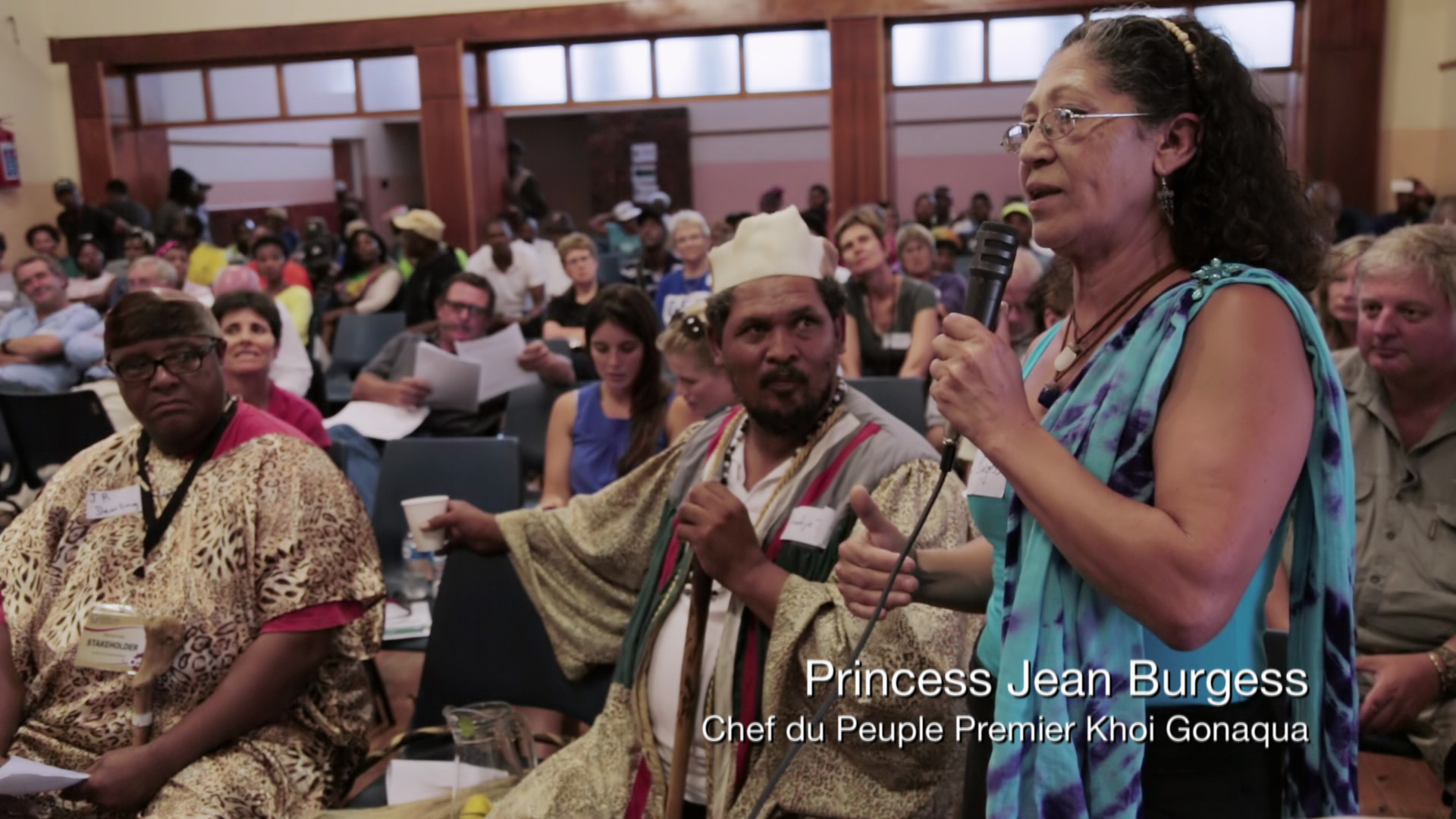 A woman in a blue dress stands and speaks into a microphone in a crowded room, with another microphone in her left hand. Seated next to her is a man in traditional attire, holding a cup. People behind them appear to be engaged in the event. Text reads "Princess Jean Burgess - Chef du Peuple Premier Khoi Gonaqua.