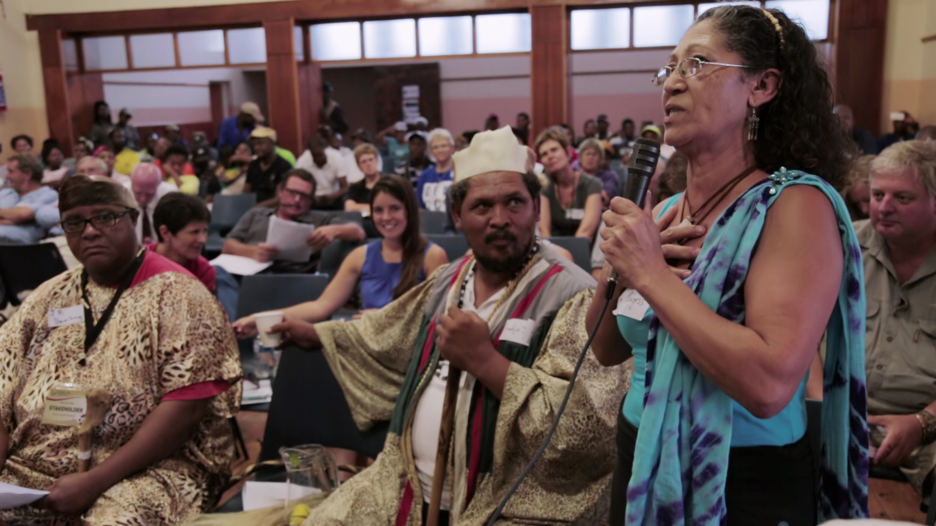 A woman in a blue dress stands speaking into a microphone in a crowded room, addressing an audience. Two men in traditional attire sit nearby, attentively listening. The room is full of seated people who are focused on the speaker.