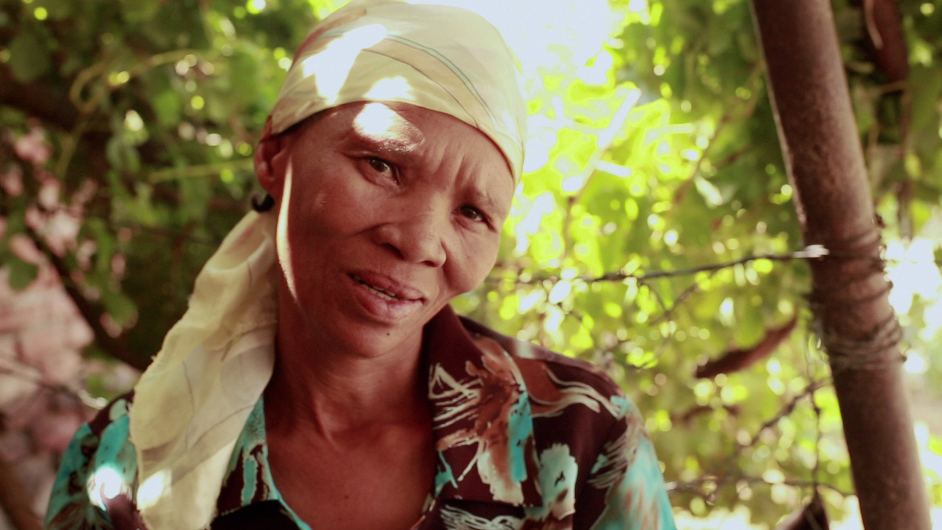 A person wearing a patterned shirt and a headscarf looks at the camera while standing outdoors under sunlight filtering through green foliage.