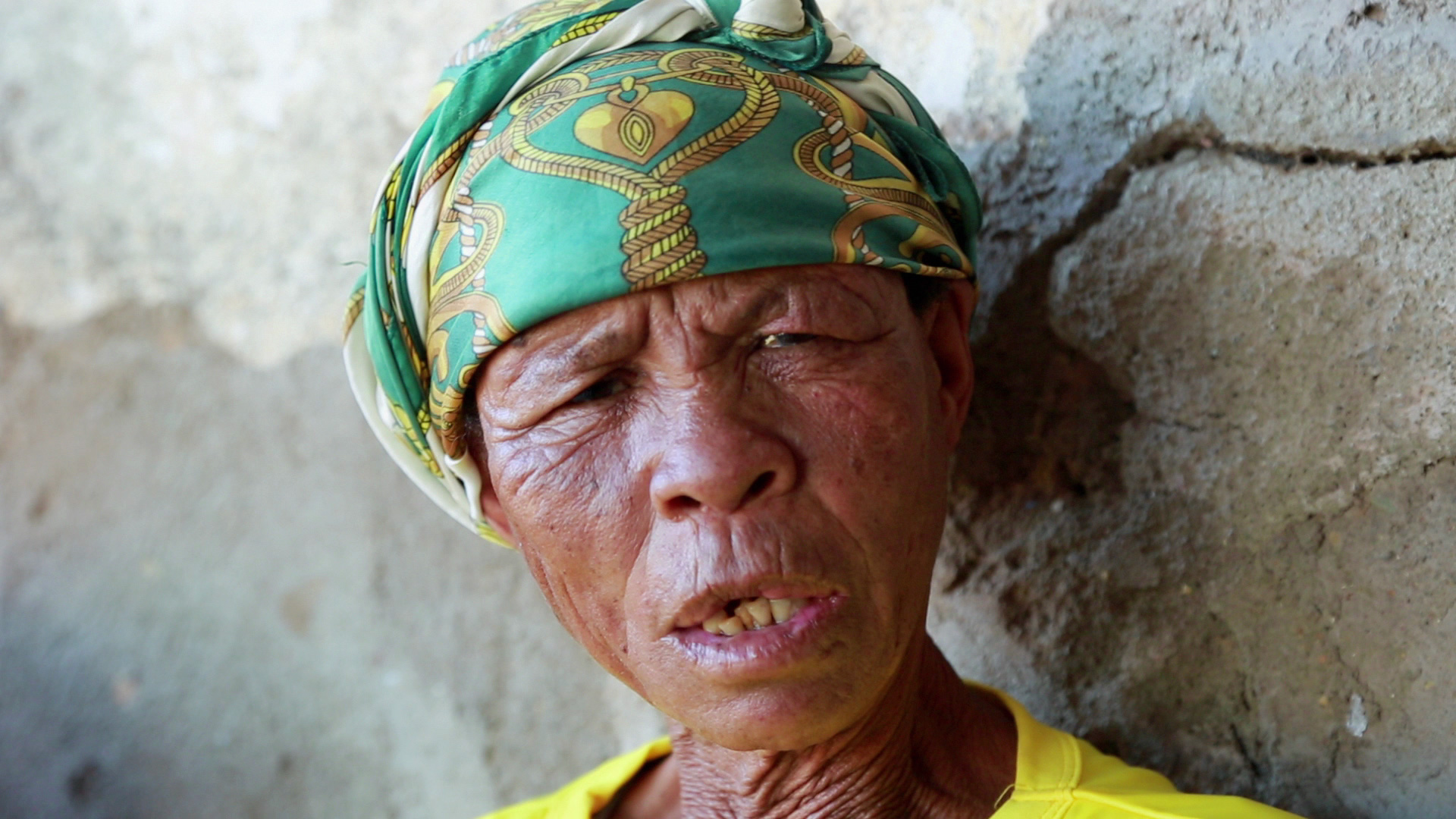 An elderly person with wrinkled skin is wearing a green and yellow headscarf and a yellow shirt, leaning against a textured wall. The person's expression is serious, with their mouth slightly open and eyes partially closed.
