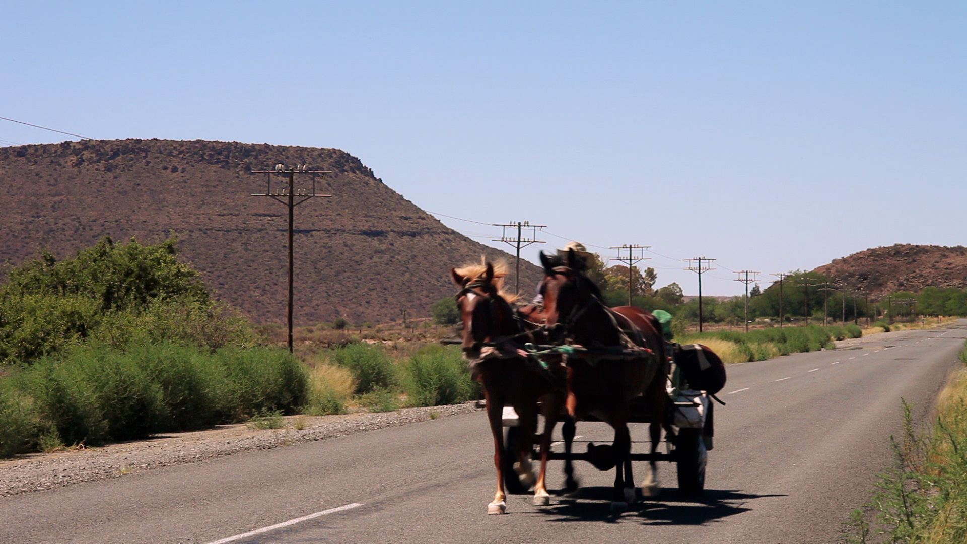 A horse-drawn carriage travels down a deserted, rural road with arid landscape and distant hills under a clear blue sky. Power lines run parallel to the road. The scene captures a blend of traditional and modern elements.