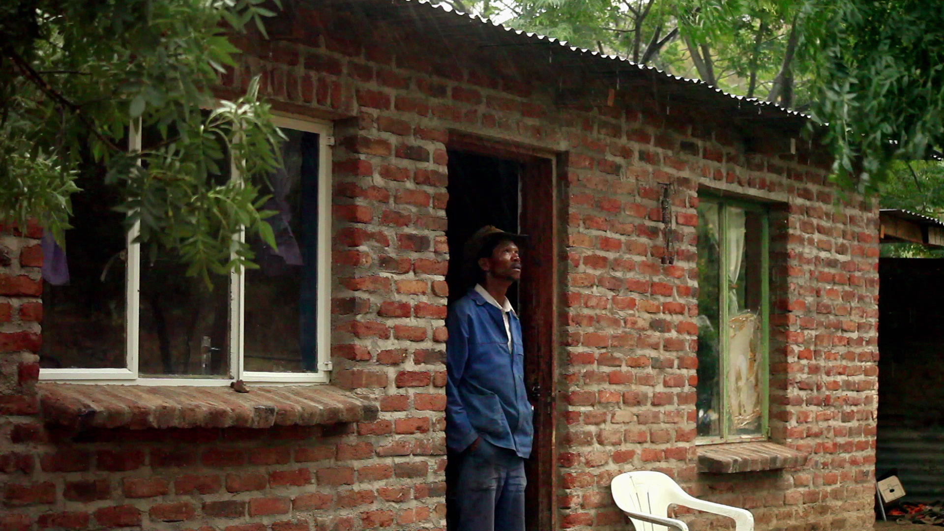 A person wearing a blue jacket and a hat stands in the doorway of a rustic red-brick house with a corrugated metal roof. The house has two windows with one partially obscured by foliage, and a white plastic chair is positioned near the entrance.