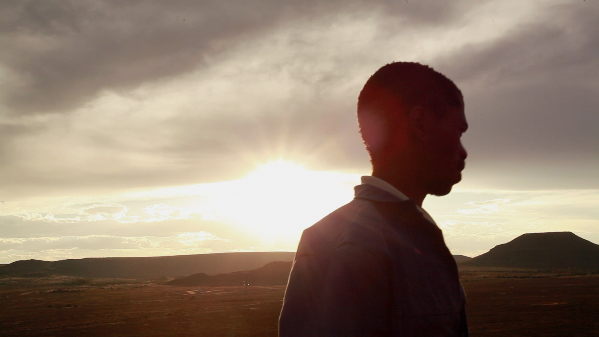Silhouette of a person standing in a vast open landscape during sunset. The sun is low on the horizon, casting a warm glow across the sky, with clouds gently dispersed. Hills or small mountains can be seen in the distant background.