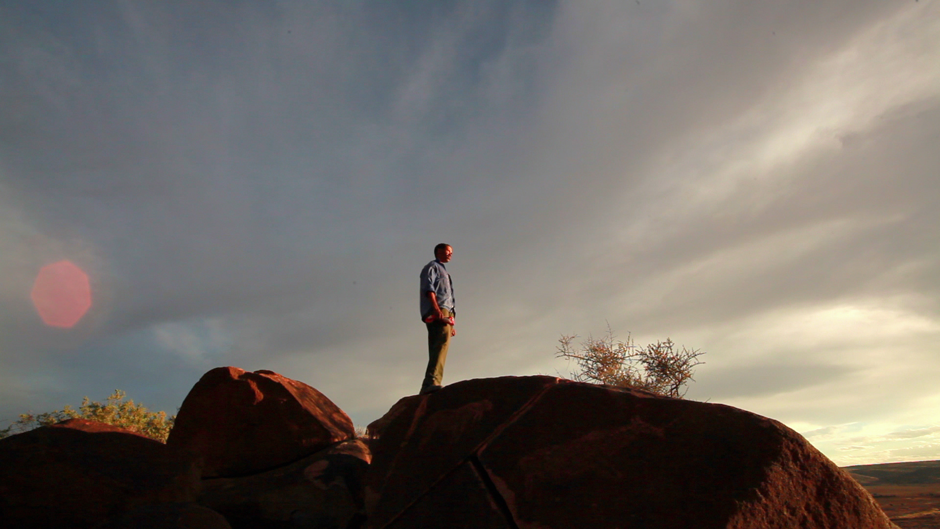 A person stands on a rocky outcrop at sunset, gazing into the distance. The sky is filled with dramatic clouds and has a golden hue from the setting sun. The individual is wearing a jacket and pants, and some sparse vegetation can be seen on the rocks.