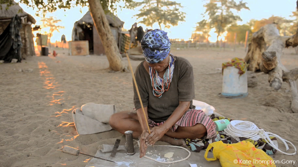 A person wearing a headscarf and colorful beaded necklaces sits on the ground in a sandy area, working with their hands. Nearby are various objects, including fabric, ropes, and bags. Huts and trees are visible in the background with a warm sunset light.