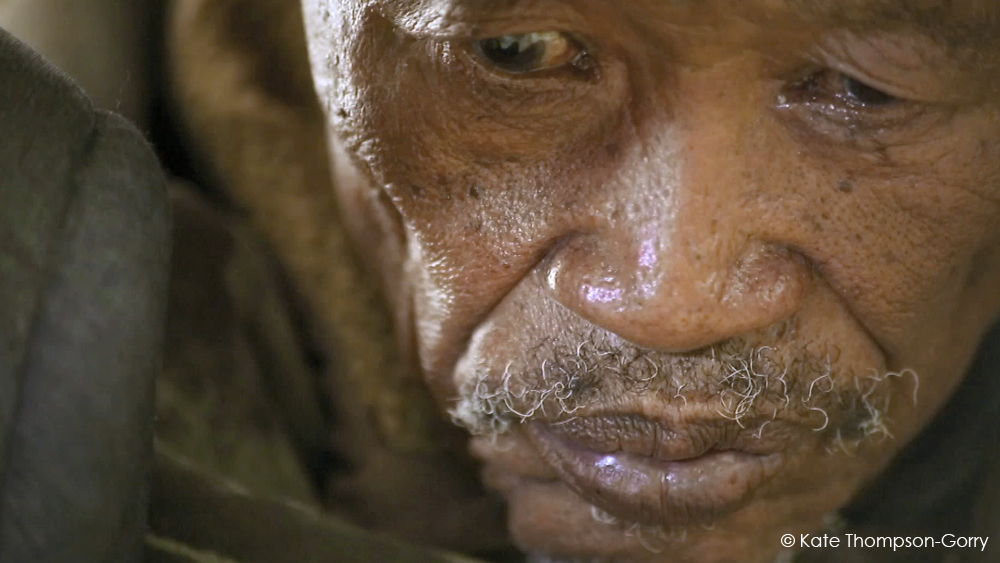 Close-up of an elderly man's face showing deep wrinkles, gray stubble, and a contemplative expression. He is looking downwards and the image captures the texture of his weathered skin. © Kate Thompson-Gorry is noted in the bottom right corner.
