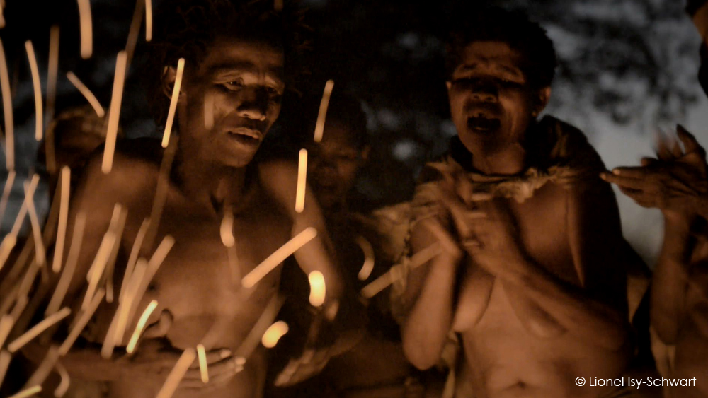 Several people are gathered closely together in the darkness with a bright fire casting sparks and illuminating their faces and bodies. They appear engaged in a communal activity, possibly singing or chanting. The atmosphere is intense and dynamic. 

Credit: Lionel Isy-Schwart.