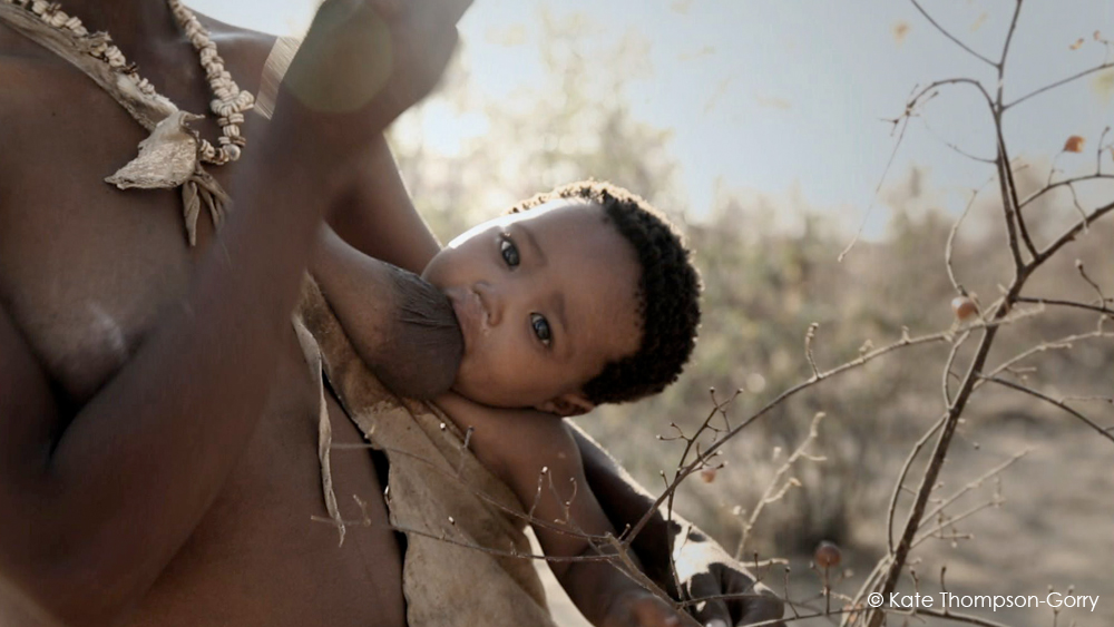 An adult holds a baby, breastfeeding outdoors. The baby looks at the camera with wide, curious eyes. The sun casts a warm glow, highlighting the baby's hair and the adult's traditional attire. Sparse foliage is visible in the background. © Kate Thompson-Gorry.
