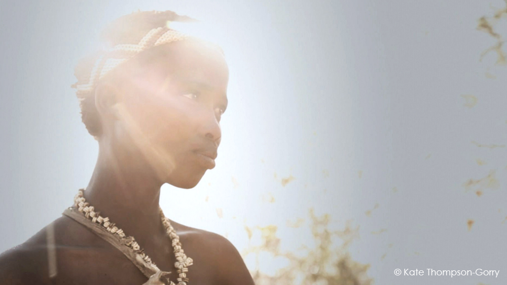 A person adorned with beads and shells is bathed in sunlight, casting a radiant glow around them. They appear thoughtful, looking to the left. The background is softly blurred. © Kate Thompson-Gorry.