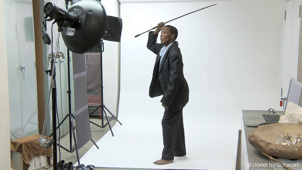 A person in a formal dark suit poses barefoot with a traditional spear, standing on a white photography backdrop. Professional studio lighting equipment and other photography tools surround the setup. © Lionel Ily-Schwart.