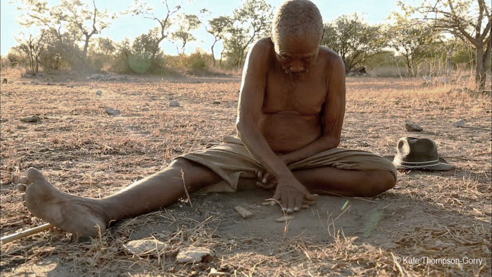An elderly person sits on the dry, cracked ground under the open sky, leaning forward while drawing in the dirt with a stick. They are shirtless and wear shorts, with a hat resting on the ground beside them. Sparse trees and a bright sky are in the background.