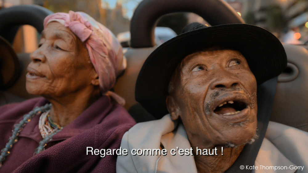 An elderly couple sitting close together, the woman on the left wearing a pink headscarf and the man on the right wearing a black hat, both looking upwards. French text at the bottom reads, "Regarde comme c'est haut !". © Katie Thompson-Gorry.