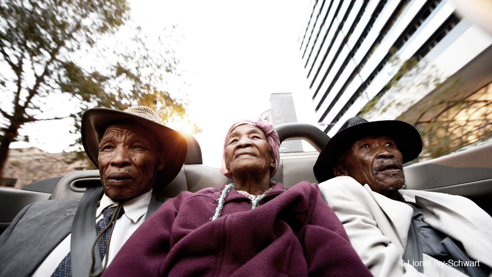 Three elderly individuals, two men and one woman, sit in the backseat of an open-top car. The woman in the middle wears a purple coat and headscarf while the men on either side wear hats and suits. Urban buildings and trees are visible in the background.