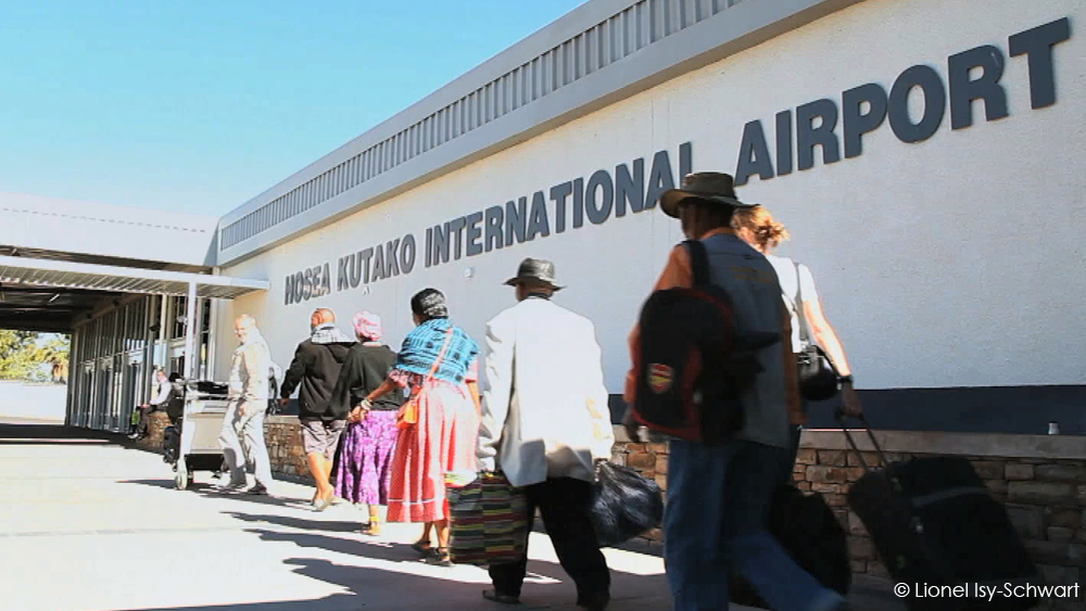 A group of travelers, some with luggage, walk toward the entrance of Hosea Kutako International Airport. The airport’s name is clearly visible on the building in large letters. The scene is bright and sunny. © Lionel Isy-Schwartz.