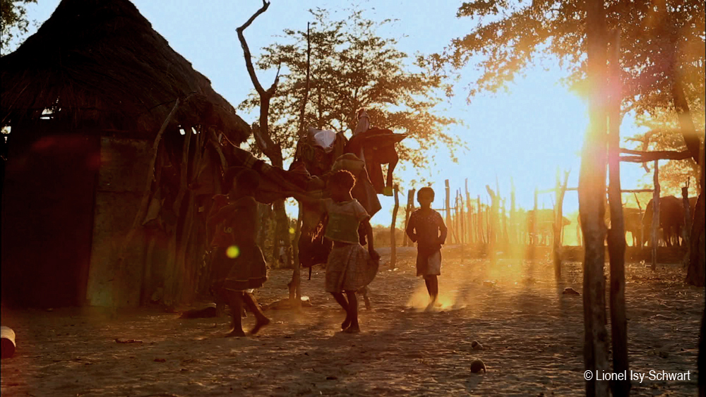 A group of children play joyfully near a thatched-roof hut at sunset. The golden light filters through trees and illuminates the dusty ground. Fencing made of sticks surrounds the area, creating a warm, serene atmosphere.