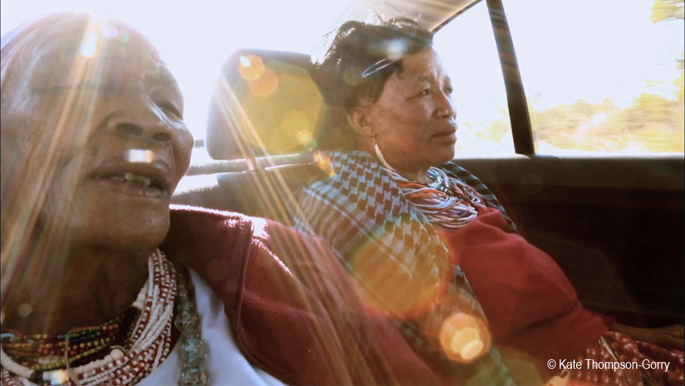 Two people sitting in the backseat of a car, bathed in sunlight streaming through the window. One is dressed in traditional clothing with beaded necklaces. Both appear relaxed and content. The image is credited to Kate Thompson-Gorry.