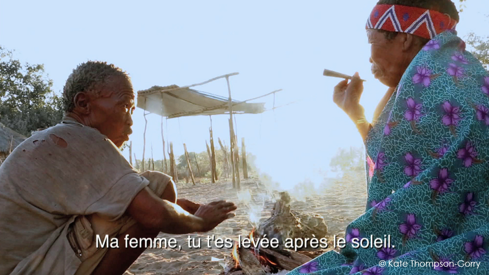 Two elderly individuals sit on the ground by a small fire. One person is wrapped in a brightly colored patterned blanket and smokes a pipe, while the other rests their hands on their knees. A makeshift shelter and trees are visible in the background. Text in French reads, "Ma femme, tu t’es levée après le soleil." The image is credited to "Kate Thompson-Gorry.