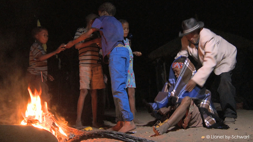 A group of children stands near a campfire, with one child helping another child stand up. An adult wearing a hat and coat is kneeling beside another person who is sitting on the ground, covered in a blanket. It is nighttime, and the scene is illuminated by the fire.
