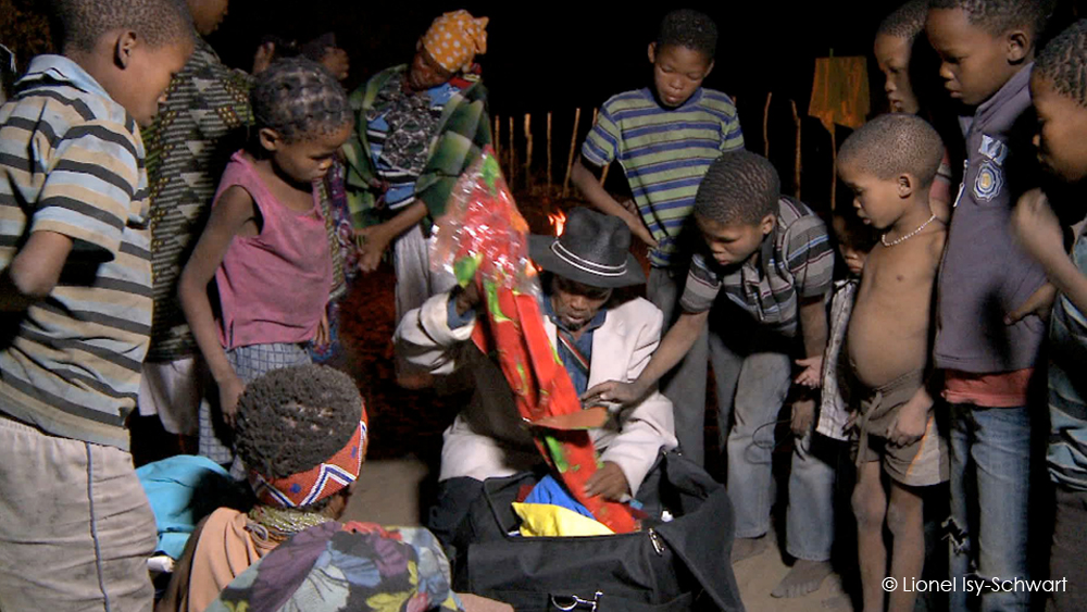 A group of children gather around an adult wearing a hat, who is sitting and holding a colorful item, possibly a gift or a toy, wrapped in plastic. The scene appears to be nighttime, with all individuals showing curiosity and interest in the activity. © Lionel Isy-Schwart is written in the bottom right corner.