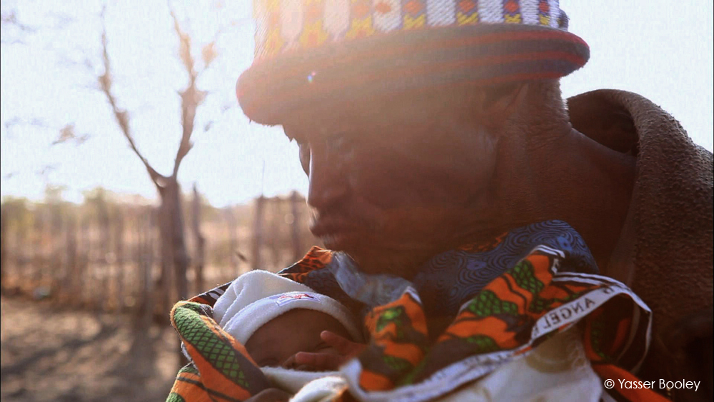 A person wearing a colorful hat holds a baby wrapped in a vibrant, patterned blanket. The scene is warmly lit by the sun, and barren trees can be seen in the blurred background. The image captures a tender moment between the two. (© Yasser Booley).
