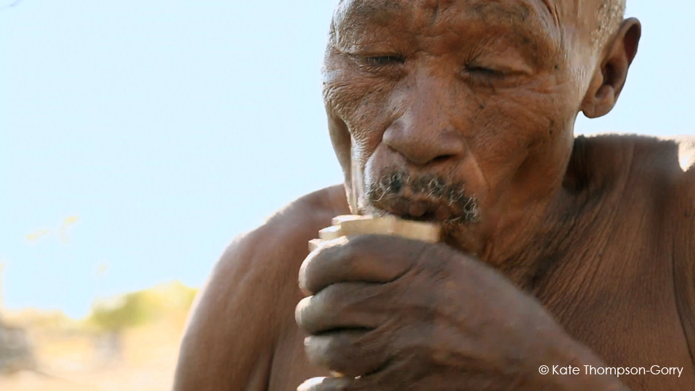 An elderly person, with visible wrinkles and partially closed eyes, is holding and drinking from a wooden cup. The background is slightly blurred, with hints of greenery. The scene appears sunlit, highlighting the person's weathered skin. (© Kate Thompson-Gorry).
