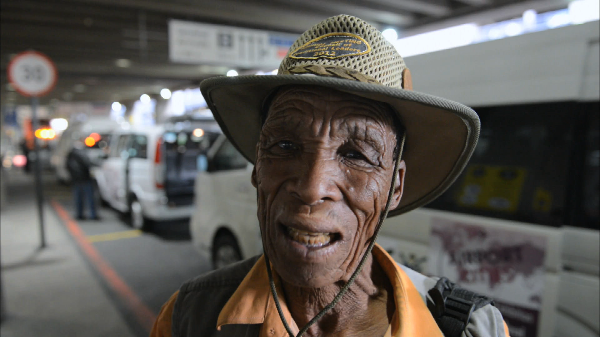 An elderly person with a weathered face and a hat stands in front of a row of vehicles in a parking area, possibly at a transport hub. The background includes a traffic sign and various vehicles, indicating a busy environment.