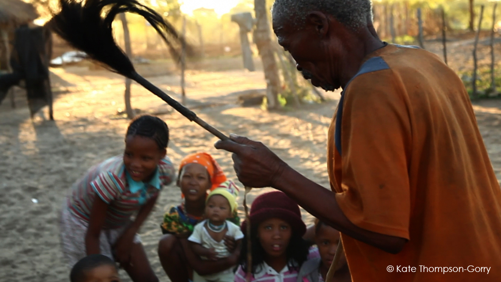 An elderly man in an orange shirt holds a feathered stick, surrounded by five children. The children, dressed in colorful clothes, look on with curiosity and excitement. The scene is set outdoors in a sandy area with trees and sunlight filtering through. © Kate Thompson-Gorry.