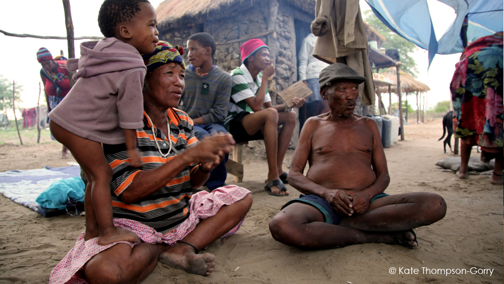 A seated group of people, including a woman with a child on her back, an elderly man with a hat, and others in the background, are gathered outdoors near rustic huts. The scene is colorful and lively, suggesting a closely-knit community. © Kate Thompson-Gorry.
